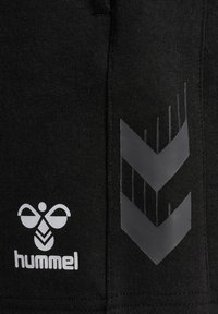 Melni sporta šorti, kas izgatavoti no mīksta auduma, ar baltu logotipu apakšējā kreisajā pusē un pelēku viegli viļņotu rakstu labajā pusē.
