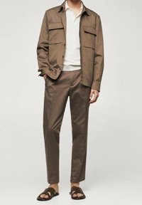 Veste légère marron avec deux poches poitrine, assortie à un pantalon assorti. Polo blanc en dessous et sandales foncées complètent le look.