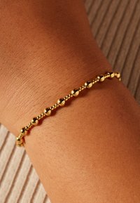 Gold bracelet featuring round beads spaced evenly along a delicate chain, resting on a tanned wrist against a textured background.
