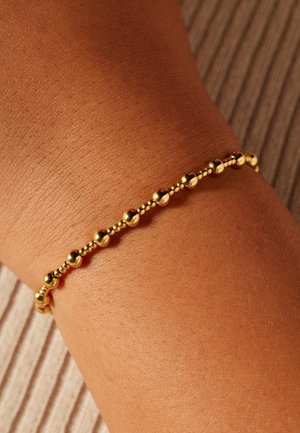 Gold bracelet featuring round beads spaced evenly along a delicate chain, resting on a tanned wrist against a textured background.