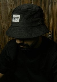 Fekete kordbársony vödörkalap, elülső címkével, ami a "Blackskies" feliratot viseli, széles peremmel és texturált anyaggal, sötét inget viselő személyen.