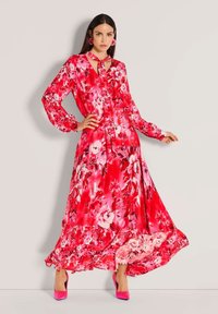 Maxikleid mit Blumenmuster in Pinktönen, mit Bindeband am Ausschnitt, langen Ärmeln und gerafftem Saum. Kombiniert mit pinkfarbenen High-Heels und passenden Ohrringen.