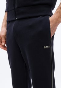 Κοντινό πλάνο ενός ατόμου που φοράει μαύρη φόρμα BOSS με το ένα χέρι στην τσέπη, με λεπτή ρίγα στο πλάι.