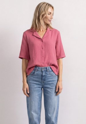 Kvinde med kort blondt hår iført en løs lyserød skjorte med knapper og halvlange ærmer samt lyseblå denimjeans, der står mod en ensfarvet baggrund.
