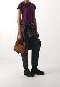 Camiseta corta de manga corta morada, camisa marrón atada a la cintura, pantalones oscuros, botas negras y un pequeño bolso de ante marrón.