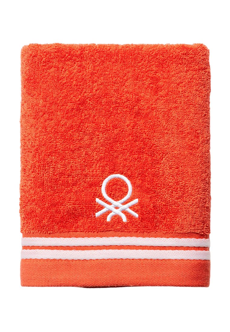 Pomarańczowy ręcznik z frotte o miękkiej fakturze. Posiada białe paski oraz okrągłe logo wyhaftowane na dole.