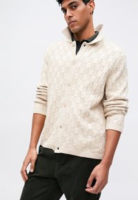 Beige gebreide cardigan met een textuurpatroon, knoopsluiting, verhoogde kraag en geribbelde manchetten, gedragen over een donker shirt en broek.