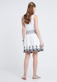 Valkoinen puuvillahame, jossa mustaa kukkakirjailua helmassa ja kuvioitu vyötärönauha. Malli pukeutuneena hihattomaan toppiin ja sandaaleihin.