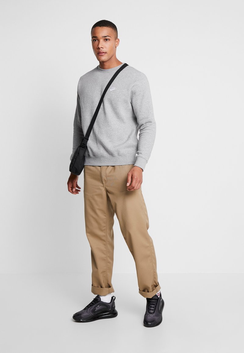 Grau Sweatshirt mit gerippten Bündchen und Saum, beige Hose und schwarze Sneakers. Das Modell trägt eine kleine schwarze Umhängetasche.