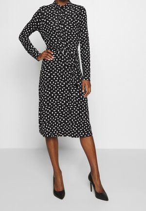 Woman wearing a black and white geometric patterned long-sleeve midi dress with a waist tie and black high-heeled shoes.