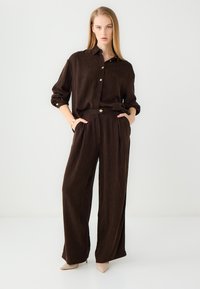 Brown linen shirt with button-up front and rolled sleeves paired with high-waisted brown linen pants. Model wears beige pointed heels.