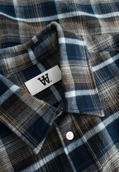 Chemise en flanelle à carreaux marron et bleu avec une étiquette blanche et un système de fermeture à boutons. Elle présente une texture douce et un col classique.