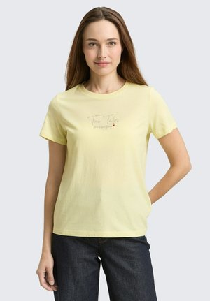 Frau mit langen braunen Haaren, die ein hellgelbes T-Shirt mit kurzen Ärmeln und dezentem Text vorne sowie eine dunkle Jeans trägt, steht vor einem schlichten Hintergrund.