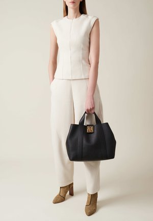 Sac en cuir noir avec un accent de logo doré, forme structurée et poignées doubles, associé à une tenue texturée de couleur crème.
