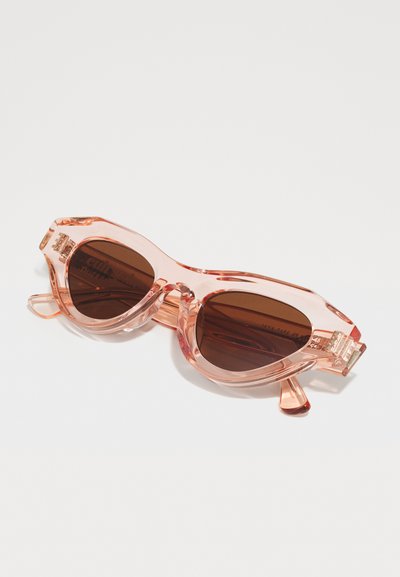 Ochelari de soare transparenți roz cu lentile maro, formă cat-eye, cadru gros și detalii accentuate pe brațe, pe un fundal alb.