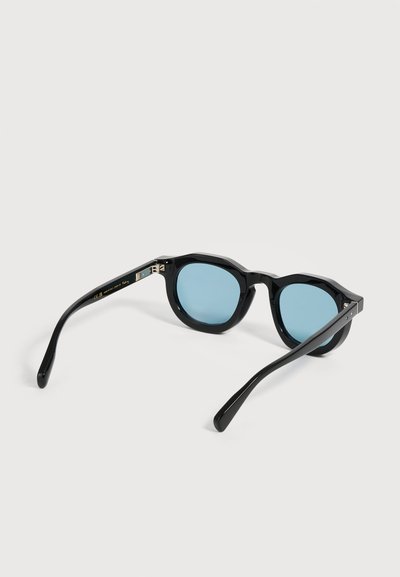 Lunettes de soleil noires à monture ronde avec verres teintés bleus reposant ouvertes sur une surface gris clair.