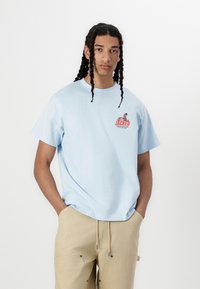 Light blue cotton t-shirt with short sleeves, featuring a colorful graphic print on the chest. Worn with beige pants, model stands confidently.