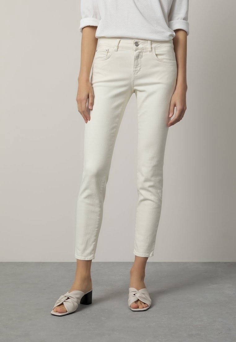 Femme portant un jean slim écru et des sandales à talons beiges ouvertes à l'avant, debout sur un sol gris sur un fond neutre.