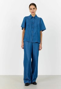Koszula jeansowa i szerokie spodnie w średnim niebieskim odcieniu. Koszula ma krótkie rękawy, spiczasty kołnierzyk oraz zapięcie na guziki z przodu.