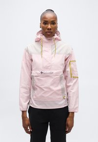 Γυναίκα με ανοιχτό ροζ και μπεζ μπουφάν Columbia anorak με μπροστινή τσέπη και κουκούλα, στέκεται σε απλό ανοιχτό φόντο.