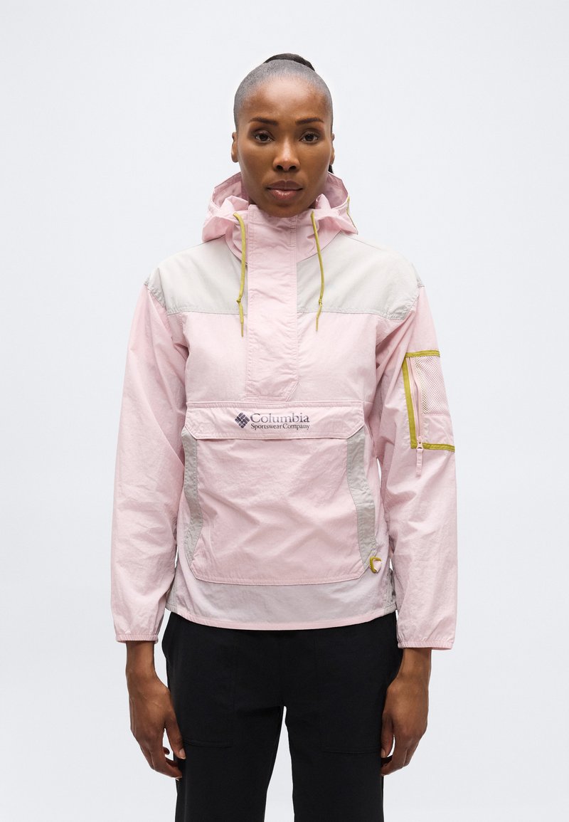 Γυναίκα με ανοιχτό ροζ και μπεζ μπουφάν Columbia anorak με μπροστινή τσέπη και κουκούλα, στέκεται σε απλό ανοιχτό φόντο.