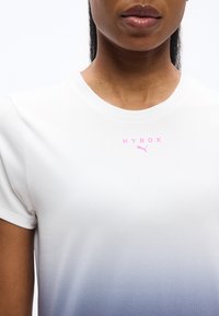 Γυναίκα που φοράει λευκό αθλητικό πουκάμισο με βαθμιαία μετάβαση σε μπλε και ροζ, με λογότυπα "HYROX" και Puma στο στήθος.