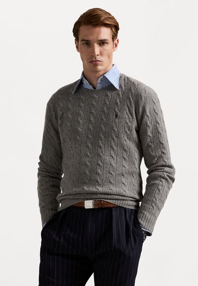Jersey de punto gris con cuello redondo, superpuesto a una camisa de cuello azul claro. Combinado con pantalones a rayas azul marino y un cinturón marrón.