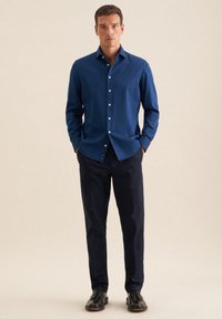 Uomo che indossa una camicia azzurra con bottoni, pantaloni blu scuro e scarpe eleganti nere, in piedi su uno sfondo beige semplice.