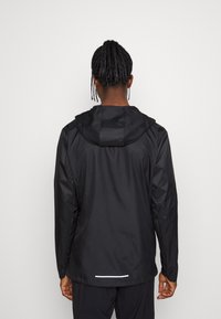 Person med flätat hår som bär en svart huva-jacket sedd bakifrån mot en plain ljus bakgrund.