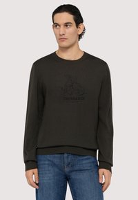 Mörkgrön tröja med rund halsringning, långa ärmar och ribbade muddar. Har en svart tryckt logotypdesign på framsidan. Bärs med blå jeans.