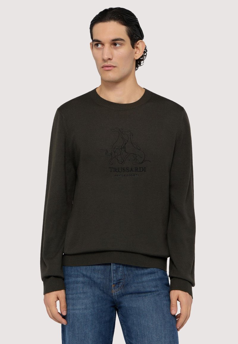 Mörkgrön tröja med rund halsringning, långa ärmar och ribbade muddar. Har en svart tryckt logotypdesign på framsidan. Bärs med blå jeans.