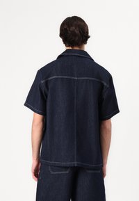 Mørkeblå denim skjorte med korte ermer og krage. Har kontrastsøm, rett snitt og detaljer på ryggen.