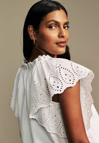 Blusa bianca con scollatura arricciata e dettagli in pizzo. Presenta motivi ricamati e un tessuto leggero e strutturato.