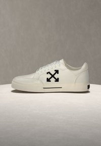 Άσπρο sneaker χαμηλού τύπου με μαύρο λογότυπο από σταυρωμένα βέλη στο πλάι, άσπρα κορδόνια και παχύ σόλα σε απλό φόντο.