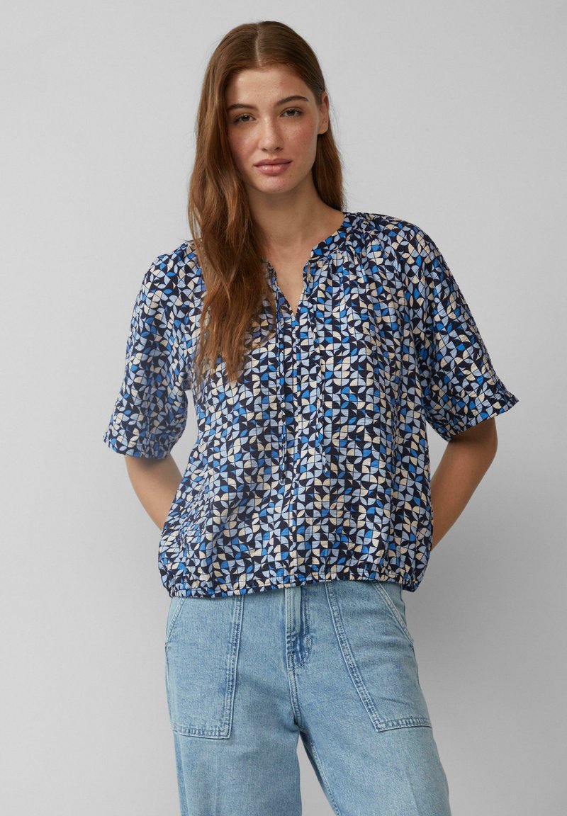 Blauwe en witte geblokte blouse met korte mouwen, een ronde halslijn en gerimpelde details aan de onderkant, gecombineerd met lichtblauwe spijkerbroek.