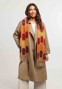 Sciarpa multicolore con forme geometriche in arancione, rosso e viola, con dettagli in frange. Indossata sopra un cappotto beige.