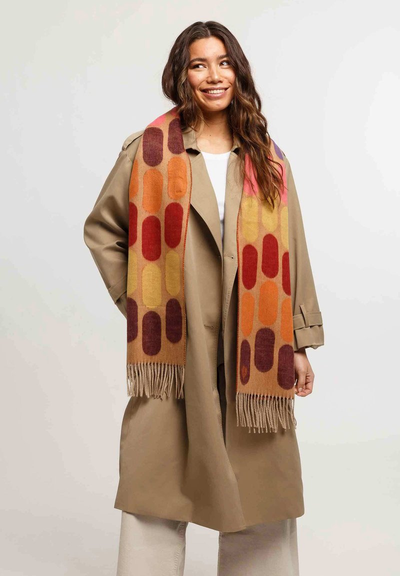 Sciarpa multicolore con forme geometriche in arancione, rosso e viola, con dettagli in frange. Indossata sopra un cappotto beige.