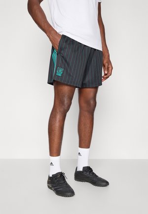 Mężczyzna w czarnych szortach Adidas z niebieskimi paskami, białych skarpetach Adidas i czarnych butach piłkarskich stoi na jednolitym tle.