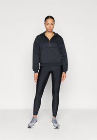 Μαύρο φούτερ με φερμουάρ μπροστά και μεγάλη τσέπη, σε συνδυασμό με γυαλιστερά μαύρα κολάν και γκρι αθλητικά παπούτσια. Απλός, εφαρμοστός σχεδιασμός.