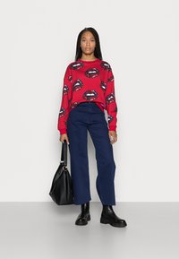 Sweatshirt vermelho com estampado repetitivo de lábios, combinado com calças largas azul-marinho. A modelo segura uma mala de couro preta e usa botas pretas chunky.