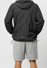 Svart lättviktsjacka med huva, elastisk fåll och dragsnören. Inkluderar "U.S. Polo Assn." logotyp på baksidan. Bärs tillsammans med grå shorts.