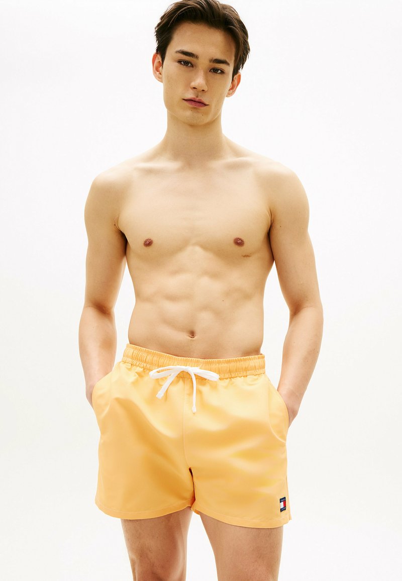 Młody mężczyzna z krótkimi ciemnymi włosami, ubrany w pomarańczowe kąpielówki z białym sznurkiem, stoi z rękami w kieszeniach na tle jasnej, jednolitej ściany.