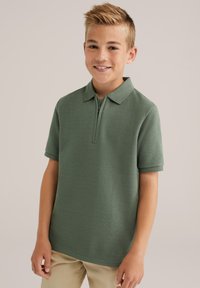 Pánské polo tričko s krátkým rukávem a zipem v texturované olivově zelené látce. Má klasický límeček a uvolněný střih. Kombinováno se světle béžovými kalhotami.