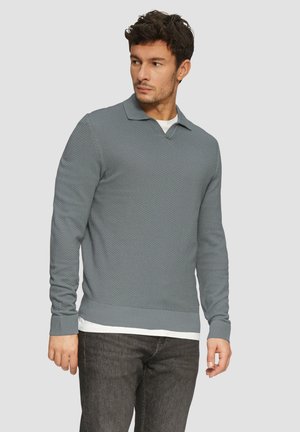Lichtgrijze gebreide trui met lange mouwen en een V-hals, over een witte shirt gedragen, gecombineerd met donkere jeans.