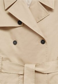 Beżowy płaszcz trench z podwójnym zapięciem, czarnymi guzikami, szerokim kołnierzem i paskiem do związania. Gładki materiał i staranne detale szycia.