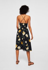 Robe florale avec un fond noir, ornée de roses jaunes et blanches. Elle a un bustier ajusté, une jupe évasée et des bretelles spaghetti.