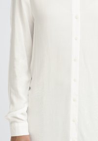 Kreis kreis un sānule valkā baltu garām piedurknēm pogājamu kreklu ar redzamām pogām un aproču detaļām.