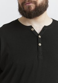 Fekete pamut henley póló, fából készült gombokkal ellátott gombos nyakkivágással, rövid ujjakkal és kerek nyakkivágással, amelynek varrott részletei vannak.