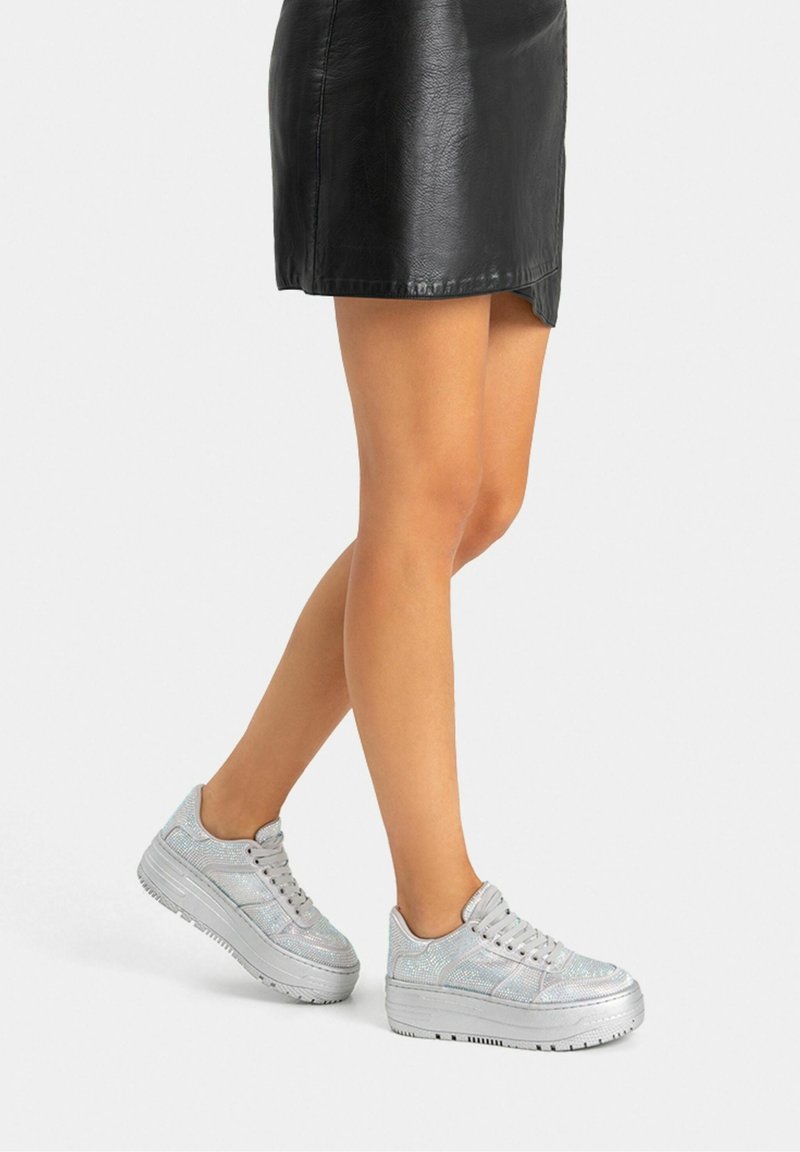 Jambes portant une mini-jupe en cuir noire et des baskets plateformes argentées sur un fond clair uni.