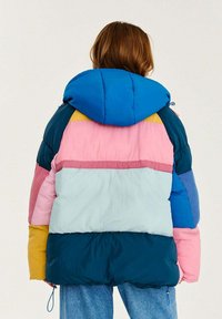 Padded jacket s kapucí, která je kombinací modrých, růžových, tyrkysových, hořčicově žlutých a tmavě zelených panelů. Texturovaný, prošívaný vzhled.